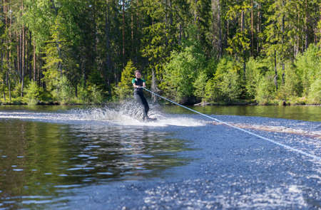 Young pretty slim brunette woman in wetsuit riding wakeboard on wave of motorboat in a summer lakeの写真素材