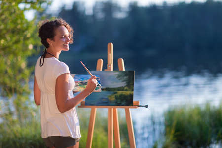 Young pretty woman artist draws paints a picture of a lake on open plain air outdoorsの写真素材