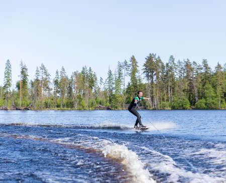 Young pretty slim brunette woman in wetsuit riding wakeboard on wave of motorboat in a summer lakeの写真素材