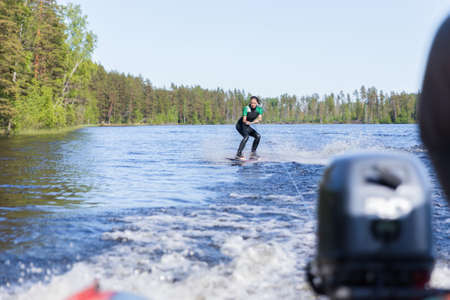 Young pretty slim brunette woman in wetsuit riding wakeboard on wave of motorboat in a summer lakeの写真素材