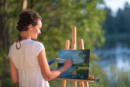 Young pretty woman artist draws paints a picture of a lake on open plain air outdoorsの写真素材
