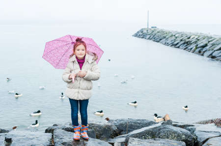 Cute little girl with pink umbrella standing next to lake on a cold weatherの写真素材