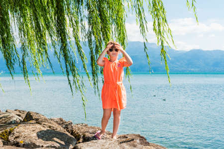 Summer portrait of a cute little girl wearing orange dress, standing next to lake on a nice sunny day, outdoorsの写真素材