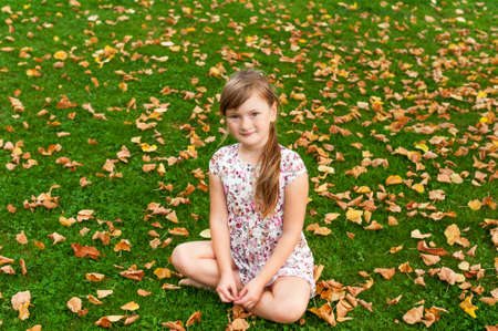Adorable little girl with long hair,  wearing white dress with flowers, sitting on grass with yellow leavesの写真素材