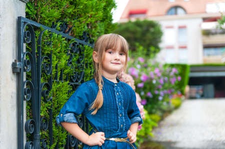 Outdoor portrait of a beautiful little girl wearing jeans dress, standing next to gate, with house on backgroundの写真素材