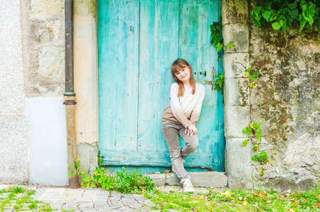 Outdoor portrait of a cute little girl, standing next to turquoise door, wearing beige pullover, pants and shoesの写真素材