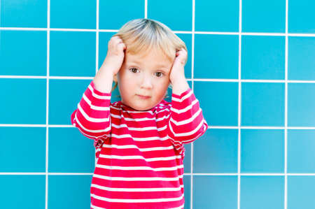 Outdoor portrait of a cute toddler boy wearing red top with white stripes, holding his head, blue tiles on backgroundの写真素材