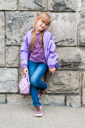 Outdoor portrait of a beautiful preschool girl wearing jeans, purple top and rain jacket, shiny tennis shoes, standing next to stone wallの写真素材