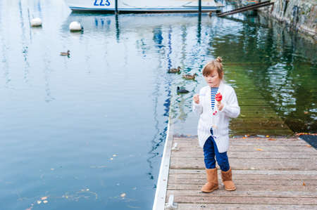 Pretty little girl wearing white knitted cardigan and warm brown boots, eating ice cream next to lakeの写真素材
