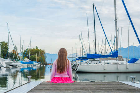 Cute little girl watching boats on the lake, sitting on a pier, view from the backの写真素材