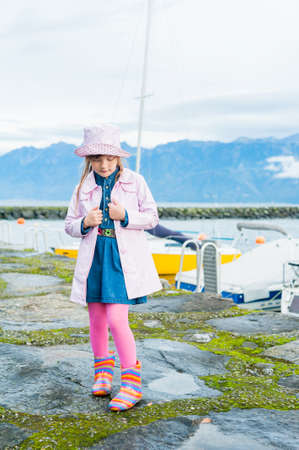 Outdoor portrait of a cute little girl, wearing pink vinyl rain coat and hat, colorful rain boots and pink tightsの写真素材