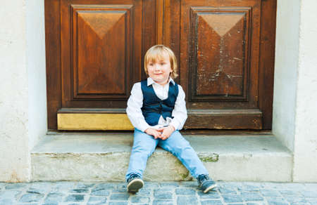 Outdoor portrait of a cute toodler boy, sitting on steps in a city, wearing beautiful celebration clothesの写真素材