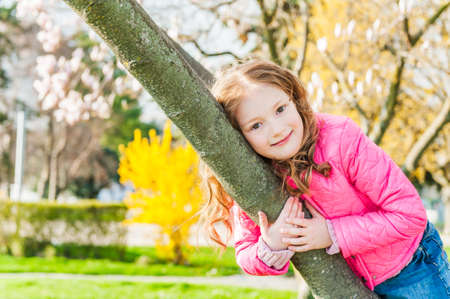 Outdoor portrait of a cute little girl in  early spring, playing on a tree, wearing bright pink jacketの写真素材