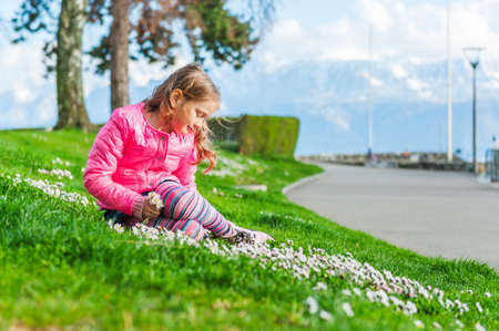 Beautiful little girl playing with flowers in a park on a nice sunny day in a early spring, wearing bright pink jacketの写真素材