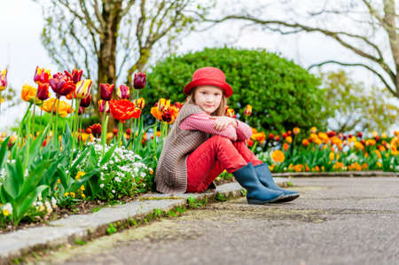 Spring portrait of a cute little girl in a red hat with red tulips outdoors in a parkの写真素材