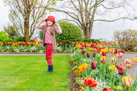 Spring portrait of a cute little girl in a red hat with red tulips outdoors in a parkの写真素材