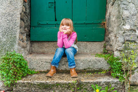 Outdoor portrait of a cute little girl, sitting on steps, wearing jeans and pink topの写真素材