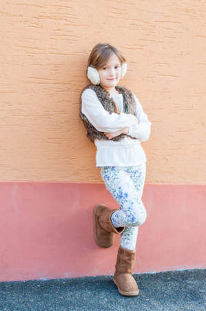 Cute little girl posing outdoors, wearing white blouse, faux fur vest, white printed jeans and...