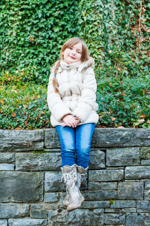 Outdoor portrait of a cute little girl sitting on a wall, wearing white warm jacket, jeans and bootsの写真素材