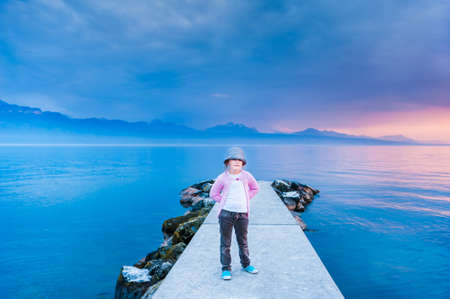 Portrait of a cute little girl on sunset standing on a pier next to beautiful lake