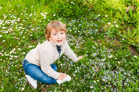 Outdoor portrait of a cute toddler boy6 sitting on a bright green grass, wearing, jeans, baskets and beige pulloverの写真素材