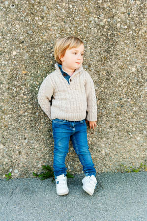 Outdoor portrait of a cute toddler boy standing next to stone wall, wearing beige pullover and jeansの写真素材