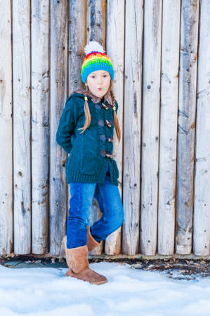 Winter portrait of a cute little girl, standing next to wooden wall, wearing, green cardigan, jeans,...