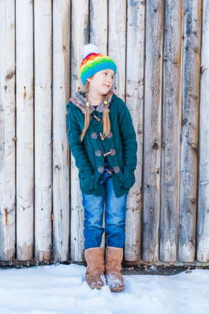 Winter portrait of a cute little girl, standing next to wooden wall, wearing, green cardigan, jeans, brown boots and colorful hatの写真素材