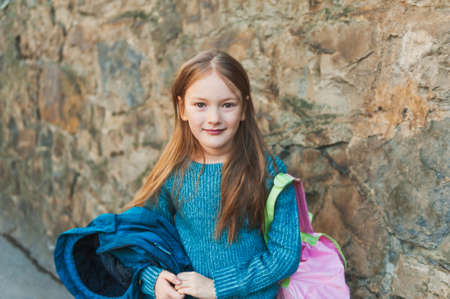 Adorable little girl of 7 years old ready to go to school, holding backpack and coat, wearing blue pulloverの写真素材