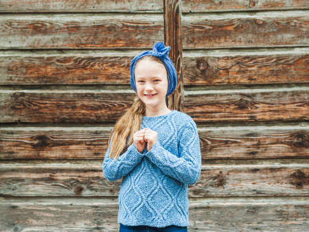 Outdoor portrait of a cute fashion little girl, wearing warm blue pullover and scarf, standing against brown wooden doorの写真素材