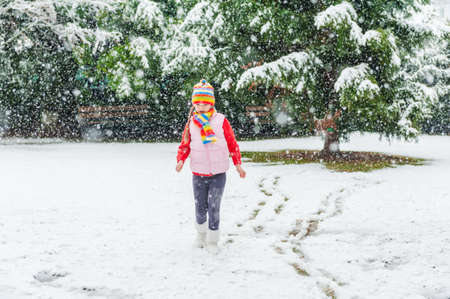 Winter portrait of a cute little girl under the snowfall, wearing red pullover, colorful hat and scarfの写真素材