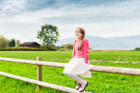 Cute little girl sitting on a fence in a countryside, wearing tutu skirt, pink pullover and bootsの写真素材