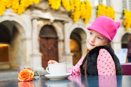 Outdoor portrait of a cute little girl drinking hot chocolate in a cafe on a nice sunny day, wearing funny pink cat hatの写真素材