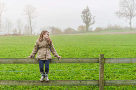 Cute little girl sitting on a fence in a countryside on a cold foggy dayの写真素材