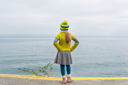 Cute little girl standing next to beautiful lake on a cold day, back view, wearing warm knit pullover and hat, bootsの写真素材