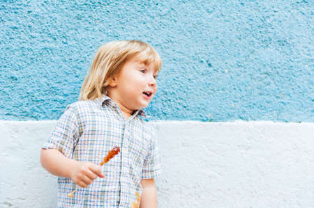 Outdoor portrait of a funny little toddler boy, holding candy, standing against blue wallの写真素材