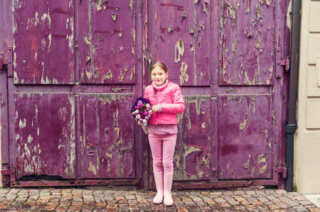 Little girl in a city, wearing bright pink clothes and boots, holding beautiful bouquet of tulips, standing next to old purple wall, toned imageの写真素材