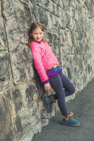 Outdoor portrait of a cute little girl on the street, wearing bright pink coat, grey leggins and...