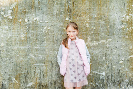 Outdoor portrait of adorable little girl of 7 years old, wearing light pink waistcoat and dress, standing against stone wallの写真素材