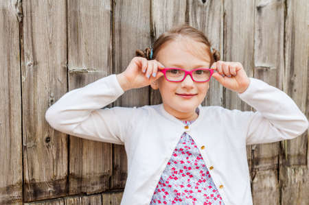 Portrait of a cute little girl of 7 years old, wearing pink eyeglasses, standing next to wooden backgroundの写真素材