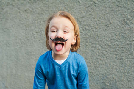 Outdoor close up portrait of a cute little boy with fake moustache, showing a tongue. Little...