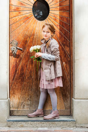 Outdoor portrait of a cute little girl of 7 years old, wearing beige coat and boots, holding white roses, standing next to old wooden doorの写真素材