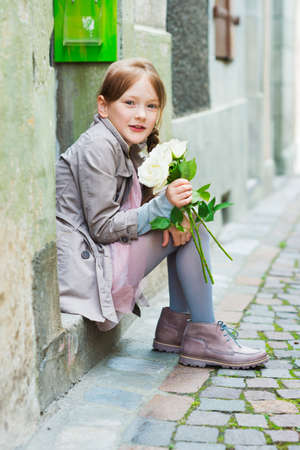 Outdoor portrait of a cute little girl with white roses, wearing beige coat and boots, sitting on steps in the old townの写真素材