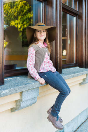 Fashion portrait of a cute little girl of 7 years old, wearing big hat, sitting by the window outdoorsの写真素材