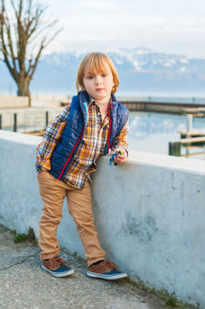 Outdoor portrait of a cute fashion little boy of 4 years old, wearing blue waistcoat, plaid shirt, beige trousers and sneakersの写真素材