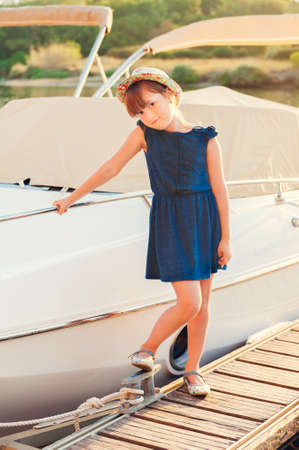 Sunset portrait of a cute little girl, standing by the lake next to white yacht, wearing blue, dress and colorful hatの写真素材