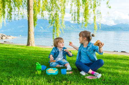 Two cute kids, little girl and her brother, having a picnic outdoors by the lakeの写真素材
