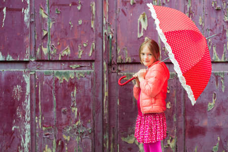 Portrait of a cute little girl next to old purple wooden door wearing pink jacket holding red umbrellaの写真素材