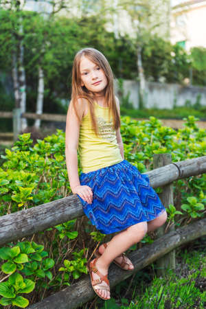 Summer portrait of a pretty little girl sitting on a fence at sunset wearing green tshirt blue skirt and brown lether sandalsの写真素材