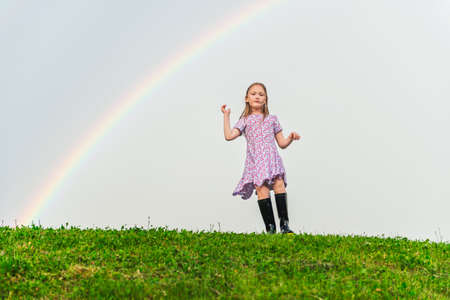 Cute little girl playing on the hill under the rainbow wearing summer dress and black rain bootsの写真素材